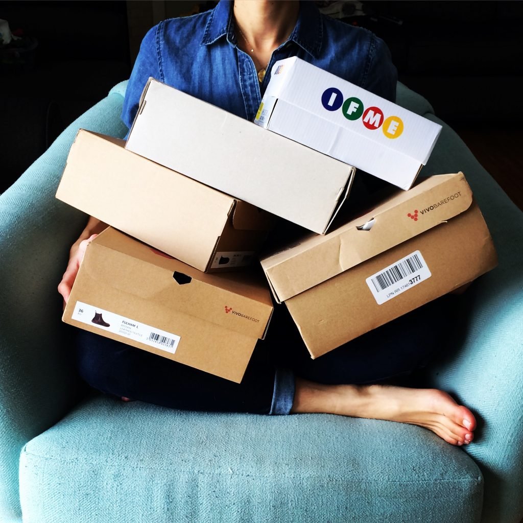 An image of a woman holding a pile of shoe boxes showing the black friday, cyber monday sales, discounts, and coupon codes on barefoot shoes and accessories.