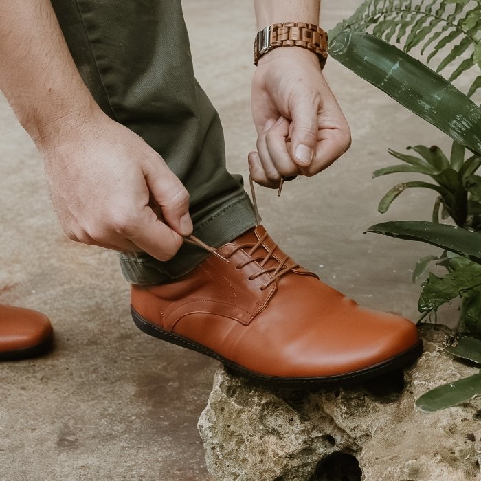 A zoomed in look at a person putting on Zaqq Peaq cognac leather barefoot dress shoes on and tying the laces
