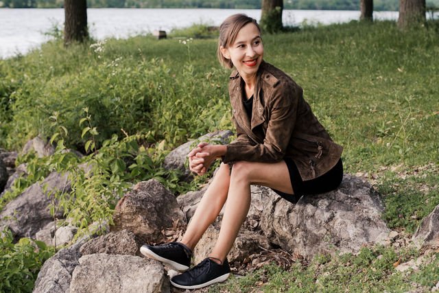 woman sitting on rocks outside smiling and looking to the side with obsidian vivobarefoot geo court sneakers on her feet and a black dress and tan leather jacket.