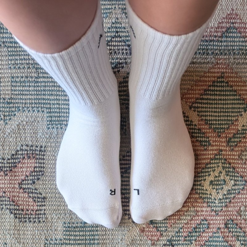 White crew anatomical socks from Aira Basics being worn by a person on a colorful rug. The L and R are stitched in black on the corresponding sock