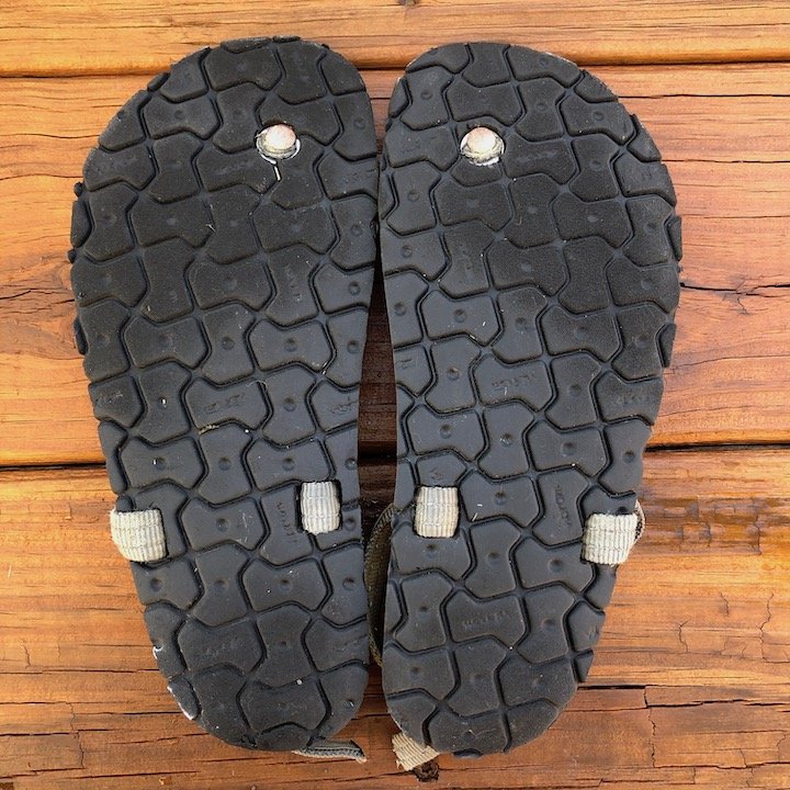 a top down view of a pair of Alpha Earth Runners sandals with a charcoal strap and cotton footbed, upside down with outsoles facing up over a wood floor