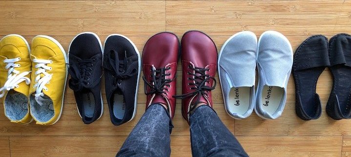 a top down view of a row of vegan barefoot shoes, with a person wearing the middle pair. Show are yellow Mukishoes, Feelgrounds Original, Red Ahinsa boots, Belenka eazy sneakers, and Oesh townie flats