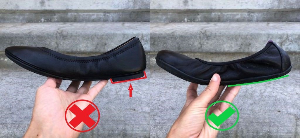 A side by side comparison of two black ballet flats being held up by a hand. On the left is a flat with a small heel (annotated with a red line) and a red X underneath. On the right is a shoe that is completely flat the whole length of the shoe with no heel whatsoever. This is annotated with a straight line and a green check mark underneath.