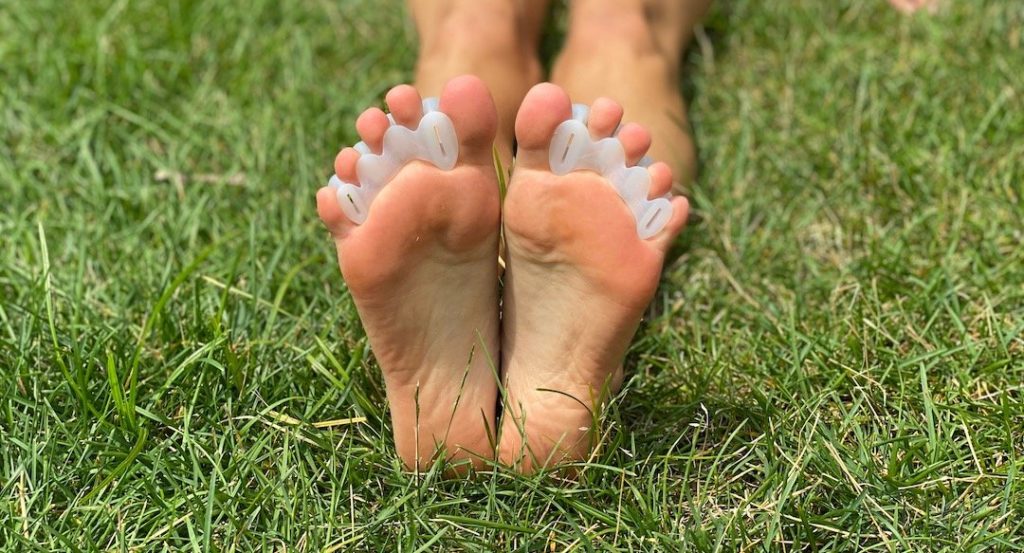 A close up of a pair of bare feet sitting on the grass with only the bottoms visible, wearing Correct Toes spacers