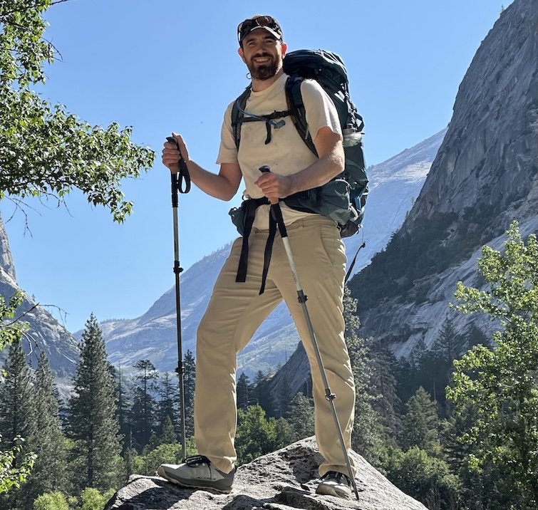 A man standing on a peak in Yosemite national park wearing a pack on his back, hiking poles, and Altra wide toe box Lone Peak hiking shoes