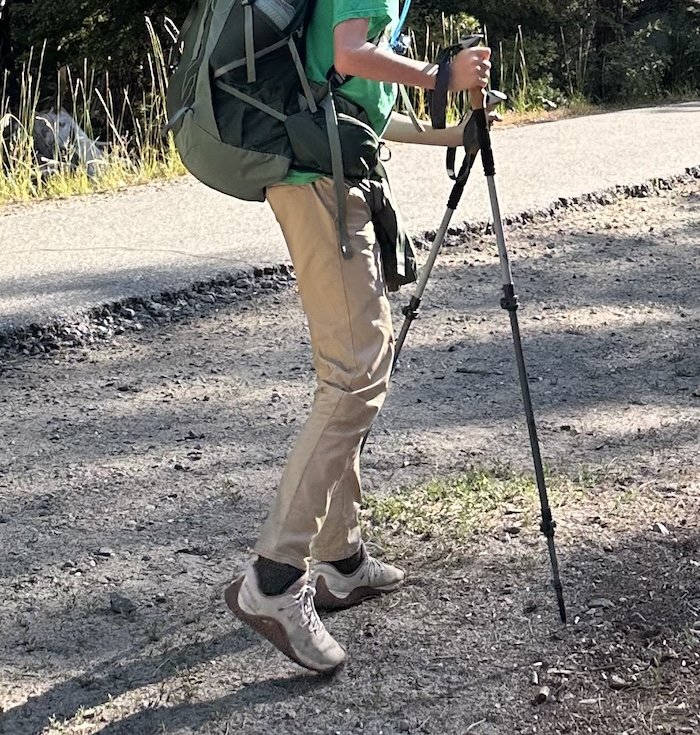 The lower half of an adolescent boy hiking with poles and a backpack wearing Merrell Vapor Trail Glove wide toe box shoes