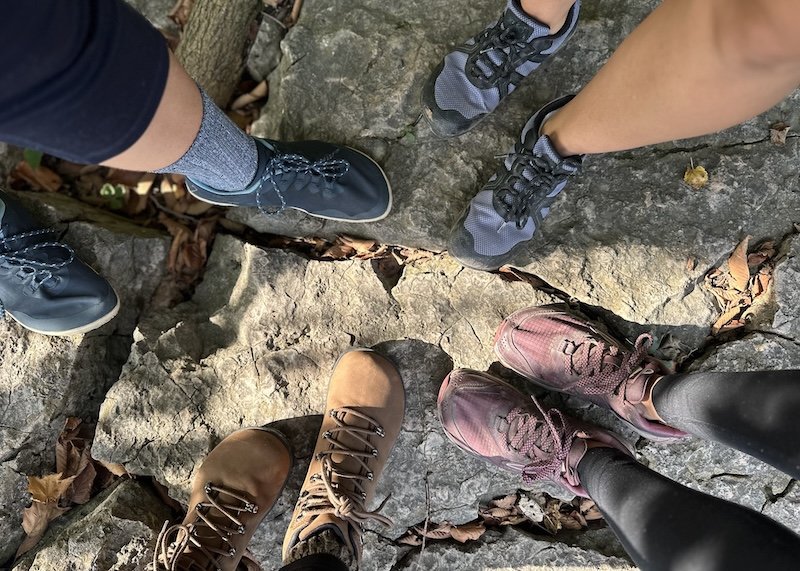 A top down view of 4 pairs of feet standing outdoors on a rockface all wearing wide toe box barefoot hiking boots and shoes