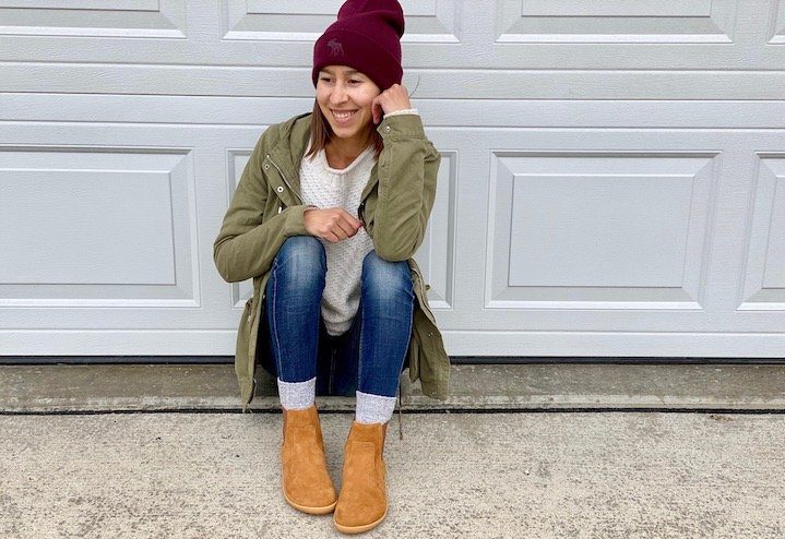 A woman sitting on the ground in front of a closed garage door wearing a coat, hat, and vivobarefoot fulham chelsea boots, smiling and looking to the side.