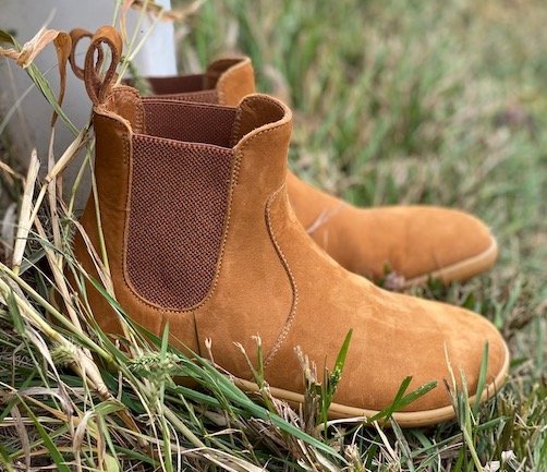 A pair of Vivobarefoot Fulham barefoot chelsea boots review in desert sand tan nubuck sitting on grass next to a white fence post, side view with the elastic panels showing.