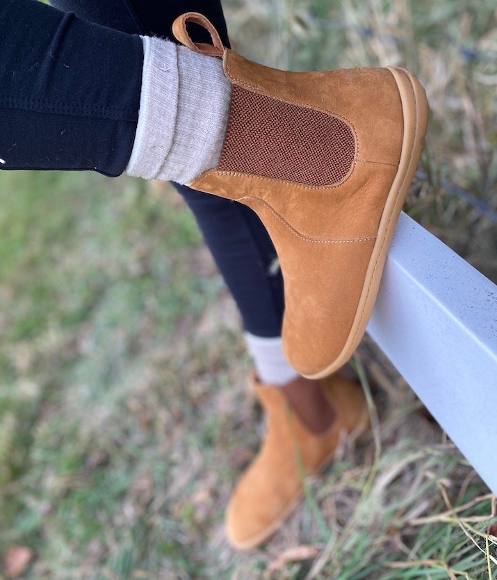 A close up of a woman's feet wearing Vivobarefoot Fulham barefoot chelsea boots in desert sand nubuck with one foot on the ground and one foot on a white fence.