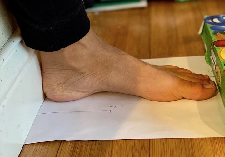 A foot with the heel linedup against the wall and a book up against the toes to show How To Measure Your Feet To Pick A Barefoot Shoe Size via the Wall Method