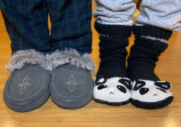 two pairs of feet standing side by side. One is wearing barefoot manitobah mukluk moccasin slippers, and the other is wearing slipper socks