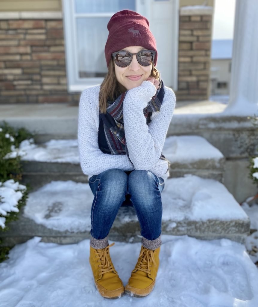 A woman sitting outside on a snowy porch smiling at the camera wearing warm clothes and Wildling shoes winter model honeybear