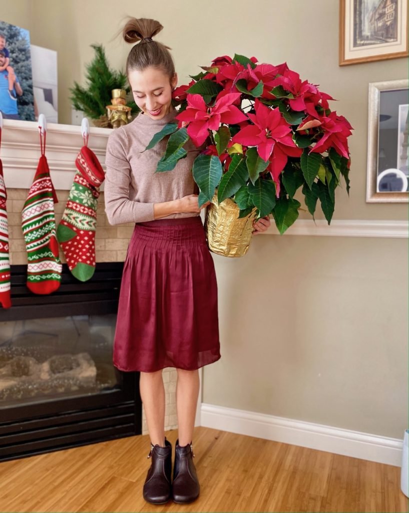 A woman dressed up in a deep red skirt, and burgundy dress ankle boots, Shapen Ivy, smiling and holding a Christmas Poinsettia