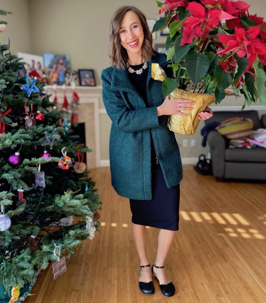 A woman holding a Christmas Poinsettia dressed in a green winter coat wearing dress shoes by Shapen, black leather Poppy flats