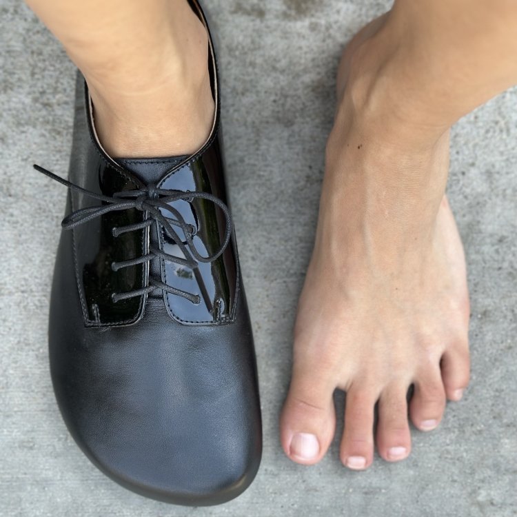 Top down close up of a woman with wide feet wearing Shapen Fleur, a barefoot oxford with patent leather accents, she has one foot bare to show the toe box width has room for toes to lay flat and prevent bunions