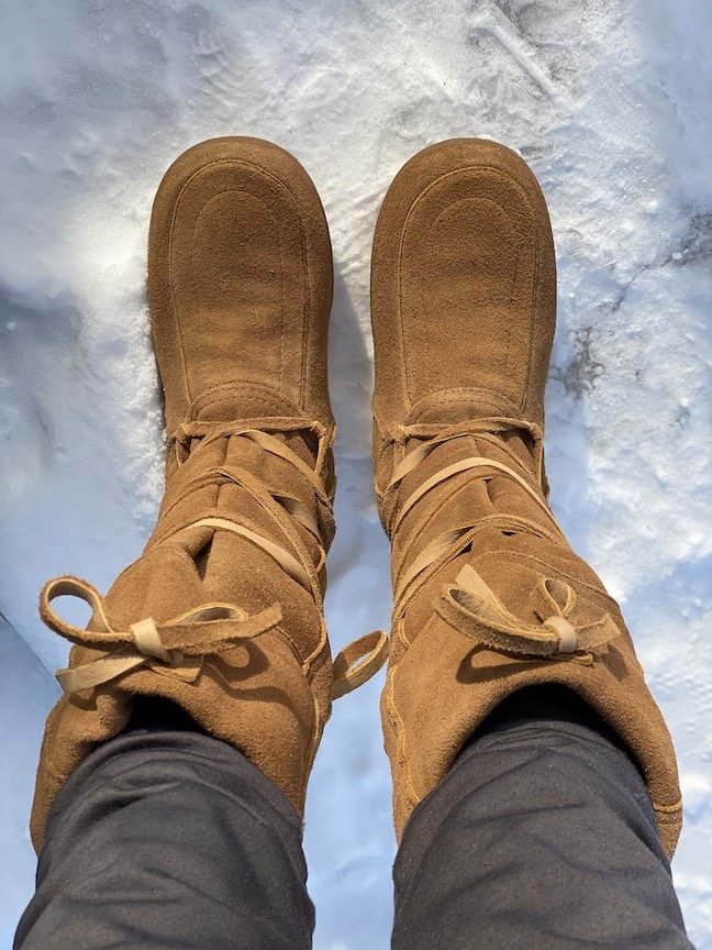 A pair of legs wearing Steger Mukluks on the snowy ground. The boots lace up to the knees.