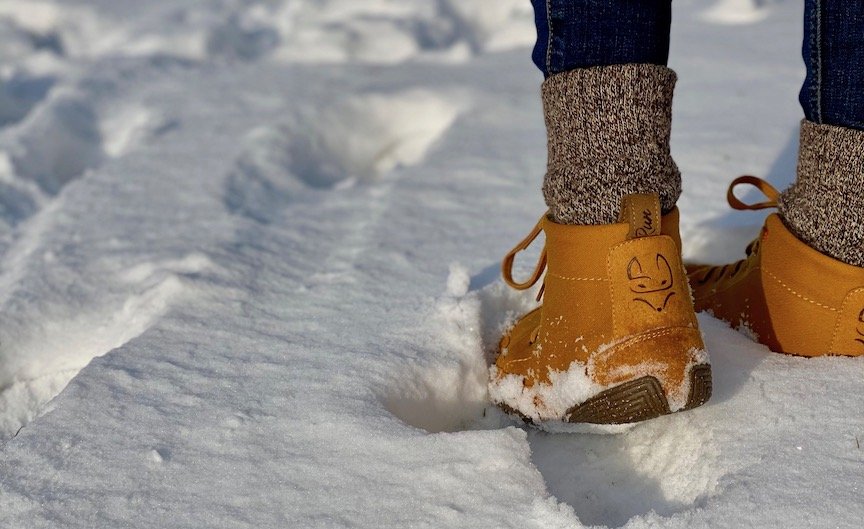 A close up image of a pair of feet from behind standing in snow that is a few inches deep. She is wearing a pair of Wildling shoes honeybear wool lined warm barefoot winter shoes.