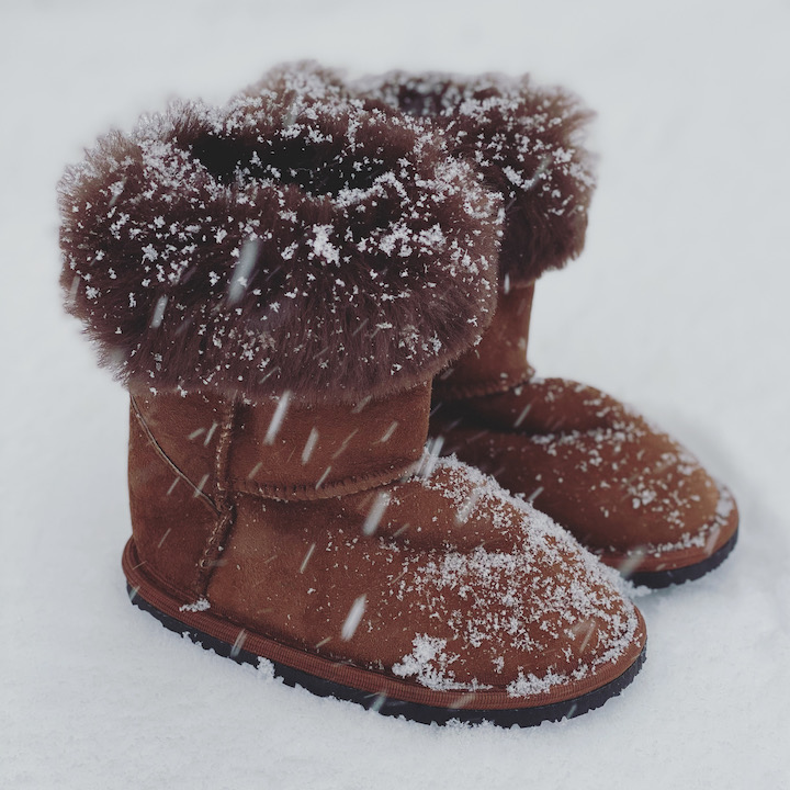 A pair of little kid Zeazoo boots sitting outside during snowfall. The sheepskin cuff is folded over and snow is gathering on the toes.