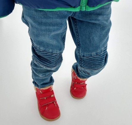 A little boy's legs standing on snow in jeans and red Ten Little High Top sneakers.