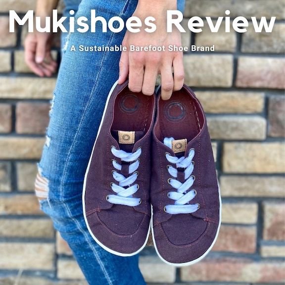 A woman holding a pair of Mukishoes Plum sneakers at her side in front of a brick wall. The text reads, "Mukishoes Review, a sustainable barefoot shoe brand."