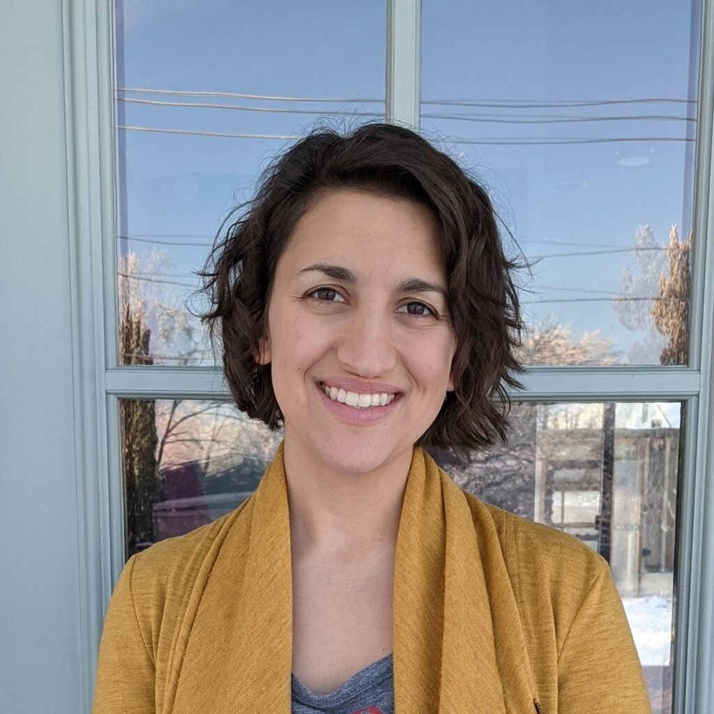 A white woman barefoot shoe blogger with brown hair smiling in front of her home.