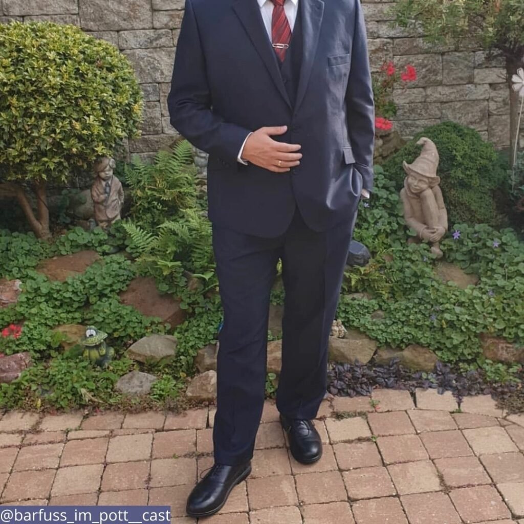 A neck down photo of a gentleman in a black suit standing outside in front of whimsical landscaping. He is wearing the best dressy barefoot shoes for men. Carets brand.