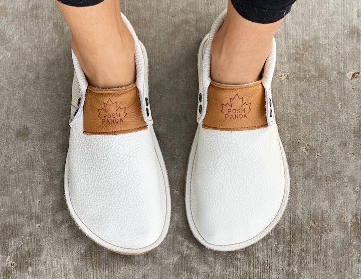 A close up of a woman's feet in a pair of white Posh Panda Moccasins. The Posh Panda logo with a maple leaf outline behind it is visible.