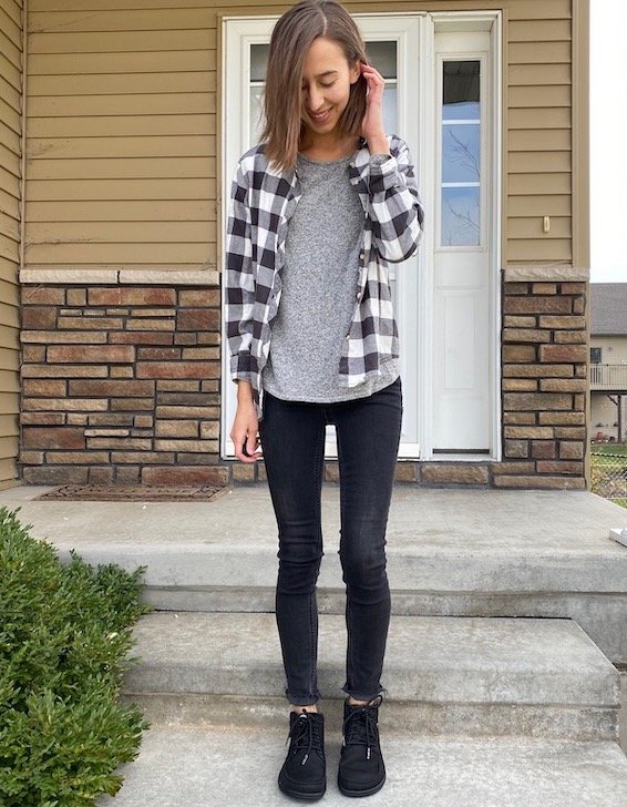 A woman standing in front of her front steps wearing black skinny jeans and a flannel button down. she is smiling down at her shoes. She is wearing Black lace up Lems Boulder Boots, a unisex barefoot option.