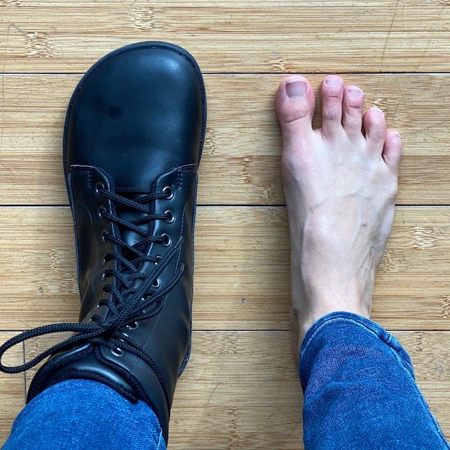 A top down view of a woman wearing one Ahinsa jaya boot standing next to her bare foot.