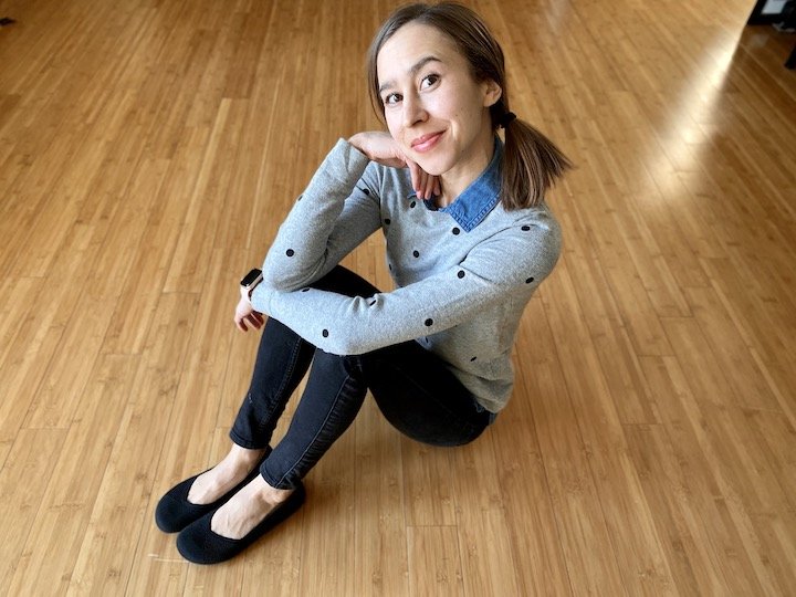 A woman sitting on a hardwood floor with her elbows on her knees making a cute face as the camera. She's wearing black skinny jeans, xero phoenix flats, and a gray sweater with sparse black polka dots.