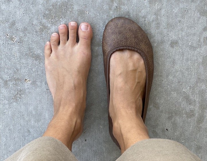 A top down view of a woman with wide feet and narrow heels wearing Softstar Ballerine flats in aged acorn on her right foot. Her left is bare to show her foot shape