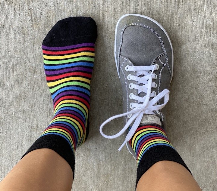 A top down view of a person's feet with rainbow socks. The left foot is shoeless, the right is wearing Splay Freestyle zero drop affordable lace up sneakers.