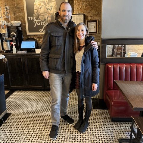 A man and woman standing with their arms around each other in a cafe wearing winter attire. The man in wearing Xero Glenn barefoot shoes and the woman is in knee high black lace up boots from Koel