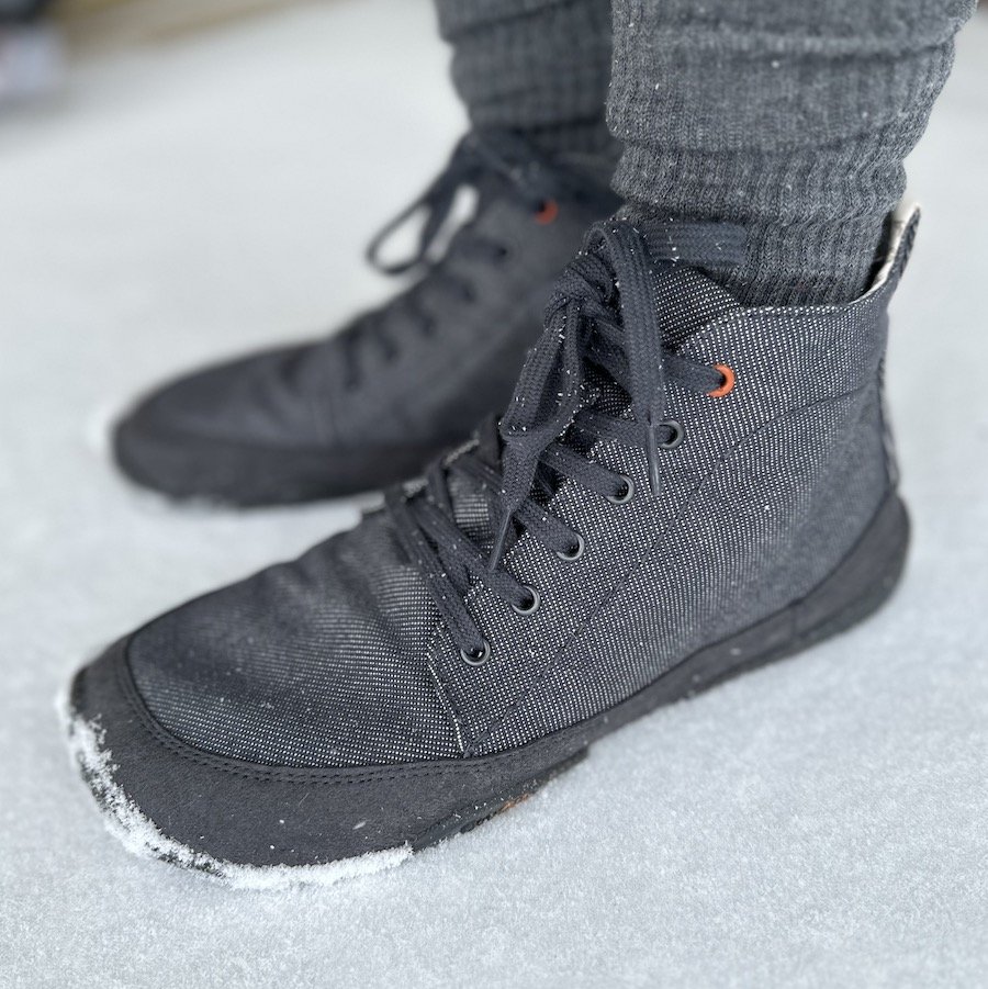 a close up side view of a pair of feet standing in snow wearing Wildling Shoes barefoot boots for fall winter in the Arni model which is made of grey-blue denim