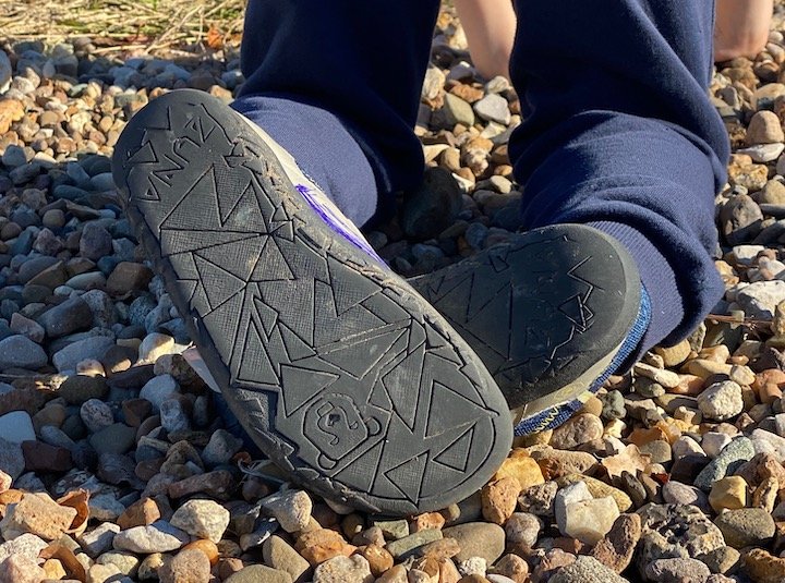 close up of a child wearing Zuna shoes barefoot zero drop sneakers while kneeling on rocks. The outsole with bear logo is facing the camera.