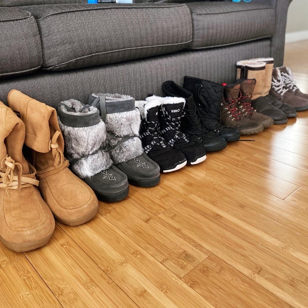 A line of barefoot winter snow boots in front of a couch showing options from Steger Mukluks, Manitobah Mukluks, Xero Shoes, Be Lenka, Vivobarefoot, and Softstar