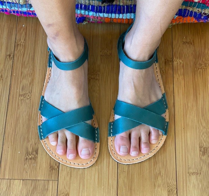 A close up view of a pair of feet wearing Crupon Barefoot Sandals in the Febo Cross Style and Avion teal leather, standing on a wood floor