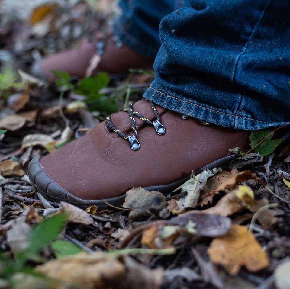 A pair of feet wearing brown vegan Freet Mudee Barefoot shoes hiking boots standing outside on dirt and leaves