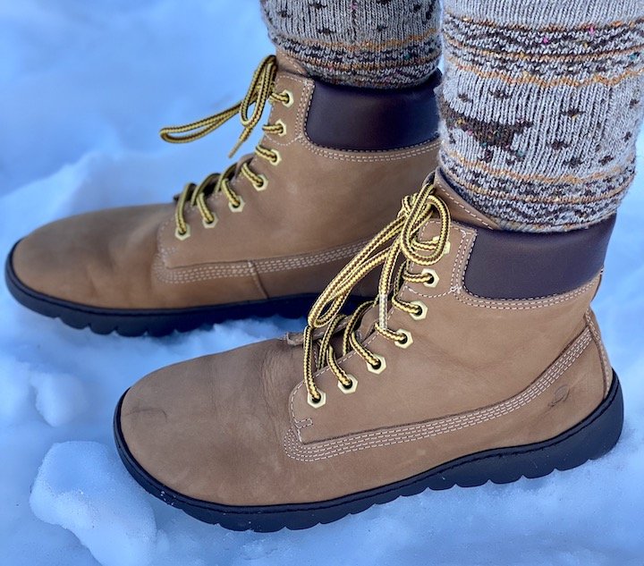 A close up of a person's feet wearing the Groundies Liverpool GX1 barefoot boot in beige leather with thick socks standing on snow side view