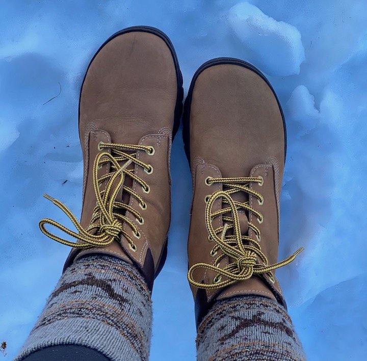 A close up of a person's feet wearing the Groundies Liverpool GX1 barefoot boot in beige leather with thick socks standing on snow top down view