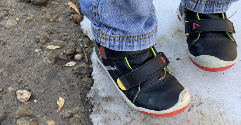 An action picture of a dark blue, red, and yellow Plae Max shoe from the top down on a frozen creek beside mud and rocks