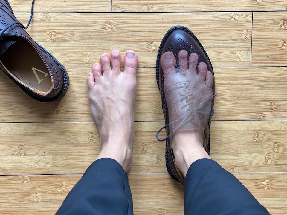 A top down view of a pair of feet. The left foot is bare and the right foot is wearing a Carets Victoria Wingtip with a superimposed translucent image of the foot inside to show that it fits without toes scrunched