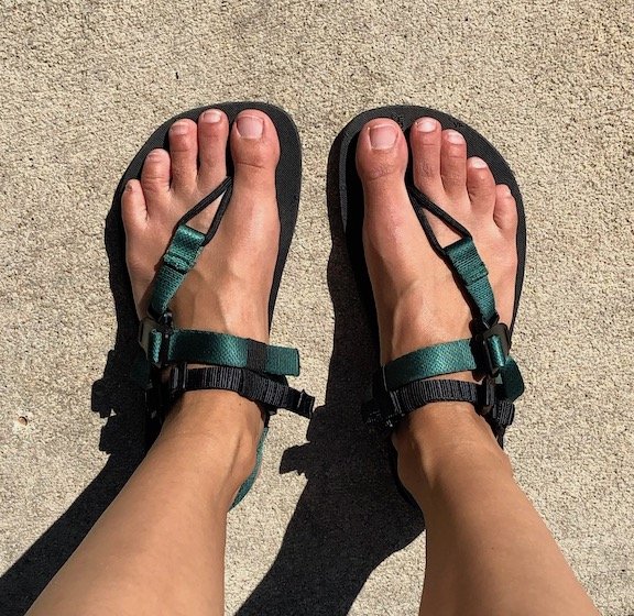 A top down view of a pair of feet standing on concrete wearing Deliberate Life Designs custom barefoot sandals in green