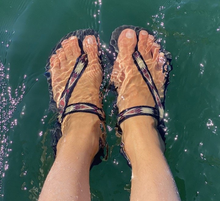 A close up view of a pair of feet wearing Earth Runners Barefoot earthing sandals in tribal print submerged in water with sunshine reflecting off