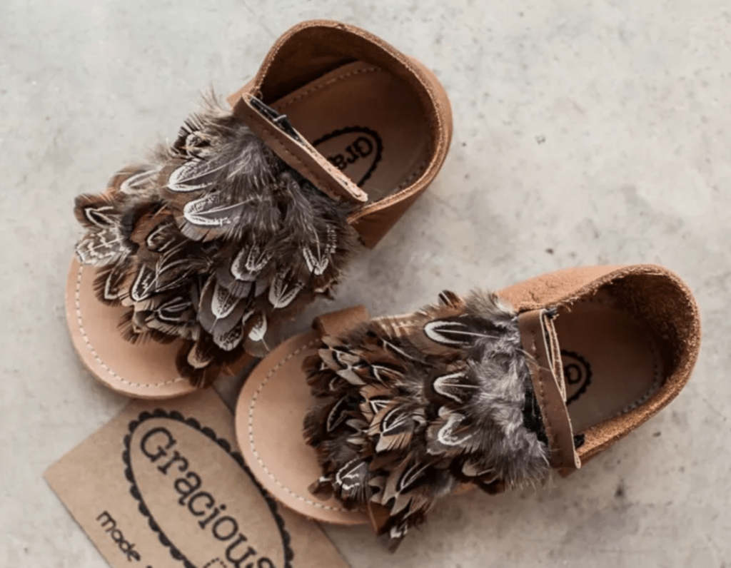A pair of brown leather barefoot sandals with feather decorations covering the top of the foot and Velcro straps shown on cement