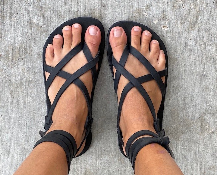 A top down view of a pair of feet standing on concrete wearing black Laboo Leather barefoot sandals