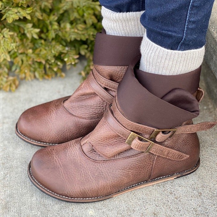 A close up side view of feet wearing the Lisbeth Joe Boulder Boot in brown leather with tan socks and jeans standing on concrete