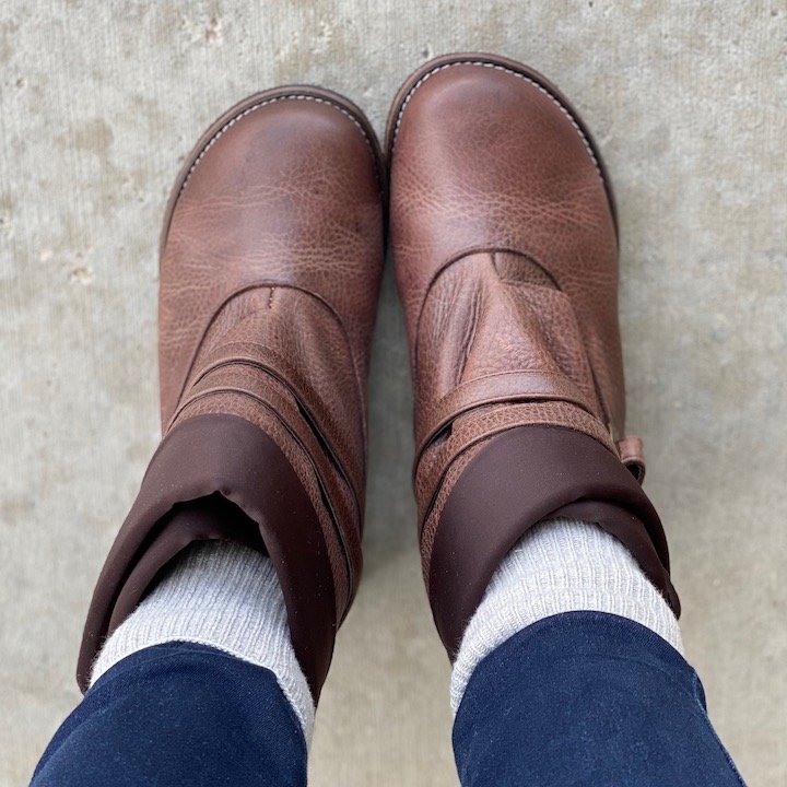 A top down view of a pair of feet standing on concrete wearing the Lisbeth Joe Boulder Boot in brown leather with the right shoe ankle strap buckled extra tight