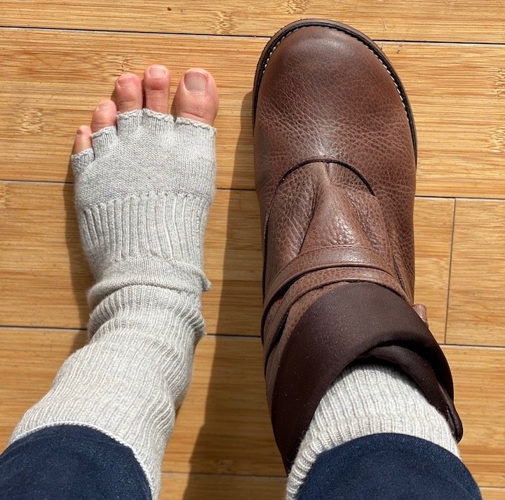 A top down view of a pair of feet standing on a wood floor - one is wearing a pair of toe socks and the other is wearing the Lisbeth Joe Boulder Boot in brown leather